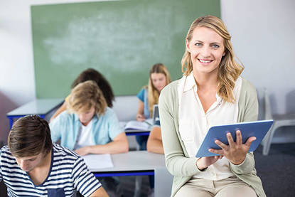 A smiling blonde teacher holds an ipad in a classroom. teenagers study at desks in the background.